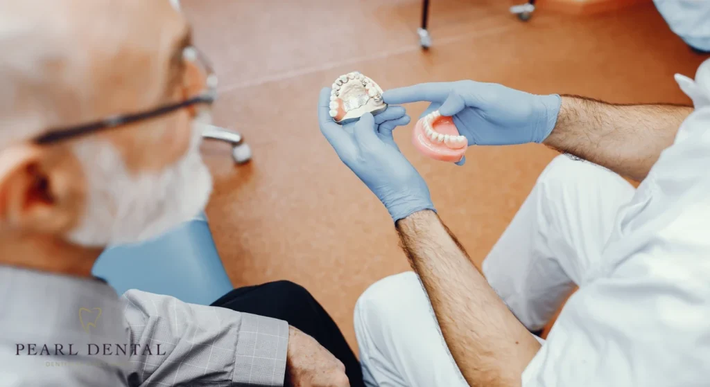 A dentist wearing blue gloves shows a model of dentures to an elderly man with gray hair. They are seated in a clinical setting, fostering a learning atmosphere.